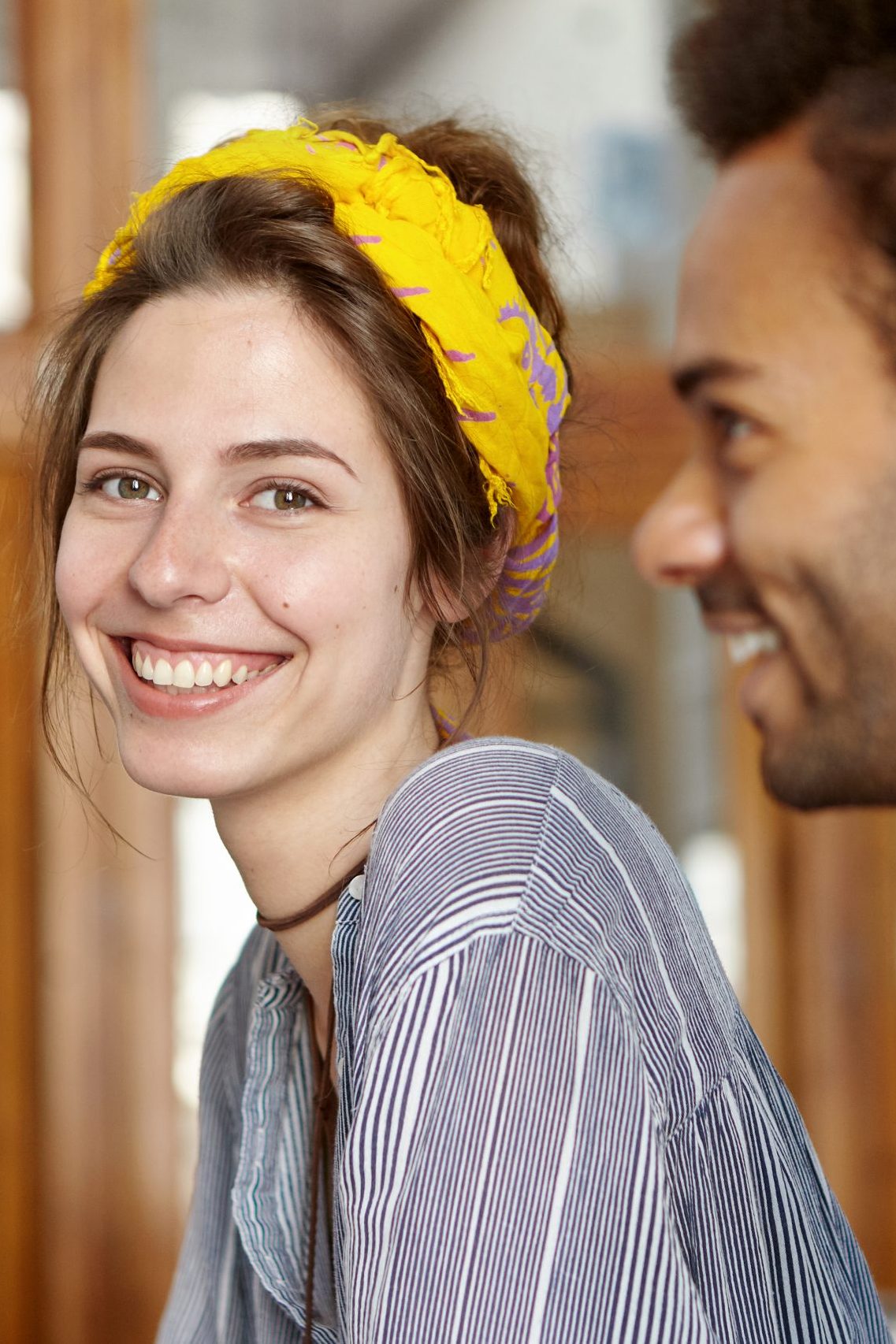 Portrait of carefree cute female wearing yellow scarf on head and striped shirt having glad expression while sitting near her African friend looking directly into camera. International friendship